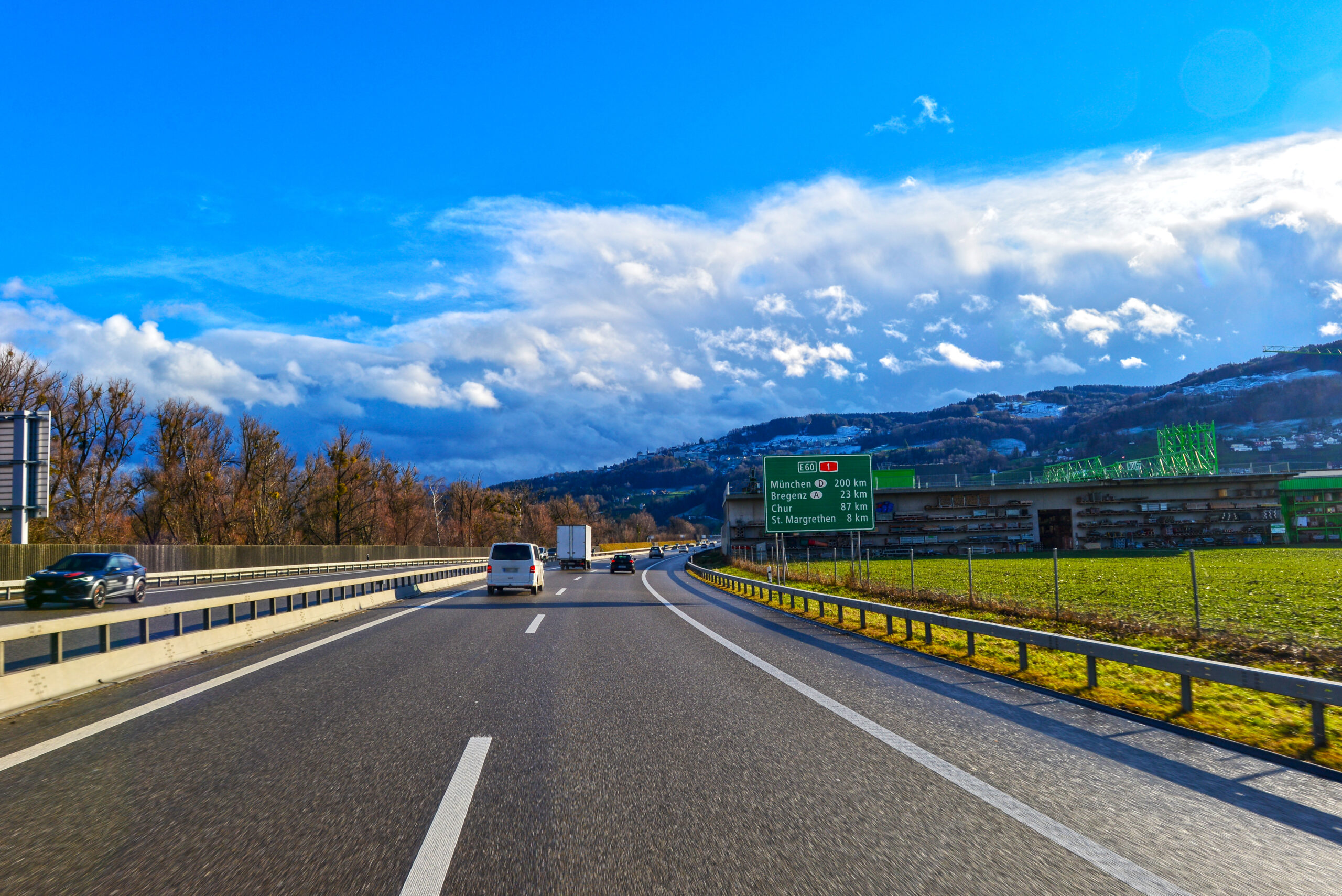 Multi-lane highway with several cars, a green road sign listing München and nearby cities, set against hills and a blue sky with clouds on a clear day.