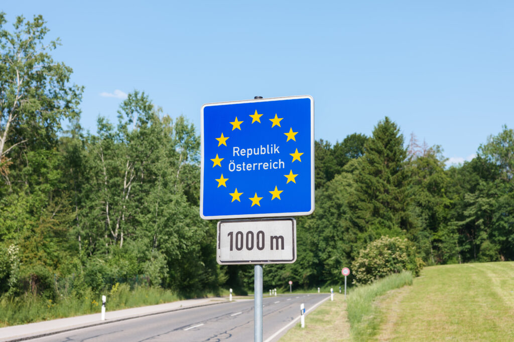 Blue road sign showing the EU circle of stars and the text ‘Republik Österreich’ to indicate entering Austria, with a 1000 m distance marker below.