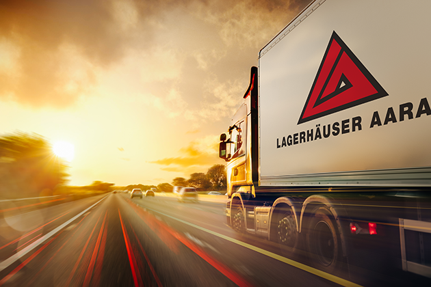 Delivery truck speeding along a sunlit highway, with motion blur in the foreground and a large trailer logo on the right.